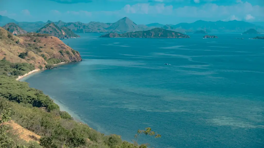 A curved bay of clear turquoise water below dry scrub-covered slopes, with island ridges on the horizon