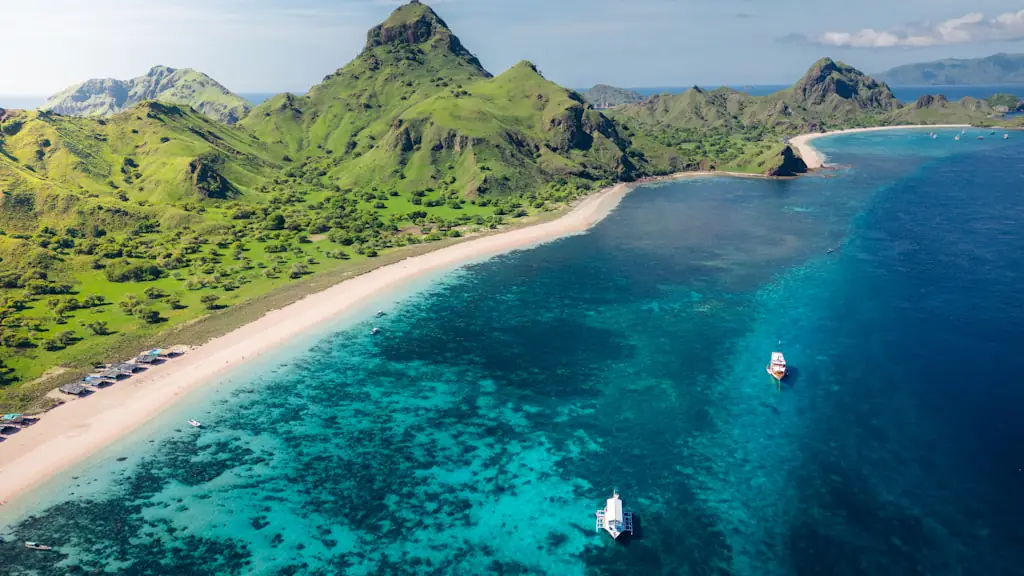 Aerial view of a green island with a curved white-sand beach, turquoise shallows and small boats at anchor