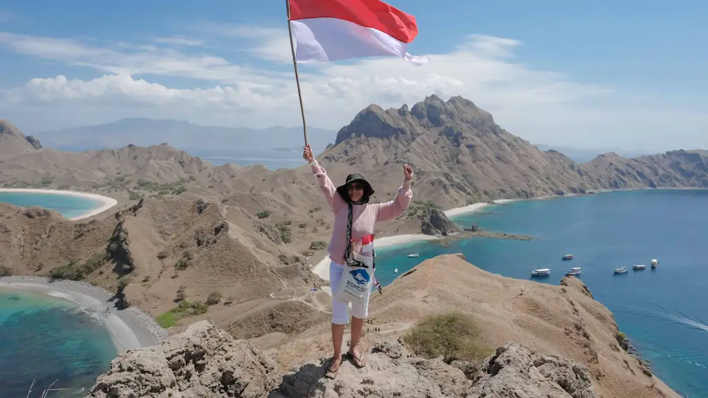 A traveller standing on a dry rocky ridge holding an Indonesian flag, with curving beaches and anchored boats in the bay below