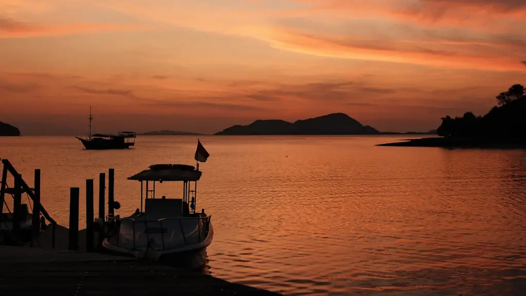 A small tour boat tied at a jetty at sunset, with a larger boat and island silhouettes on the orange water beyond