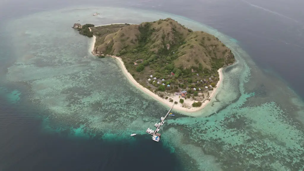 Aerial view of a large hilly island ringed by shallow reef, with a beachfront cluster of pale tent-cabins and a red-roofed building, a wooden jetty and moored boats