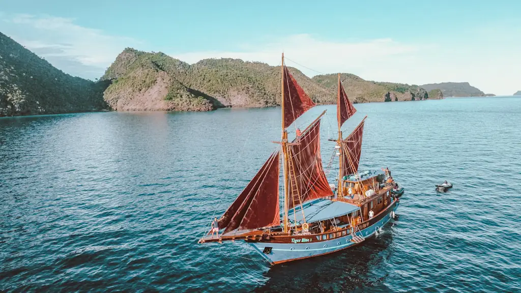 Phinisi sailing boat with red sails on calm water beside a steep green headland