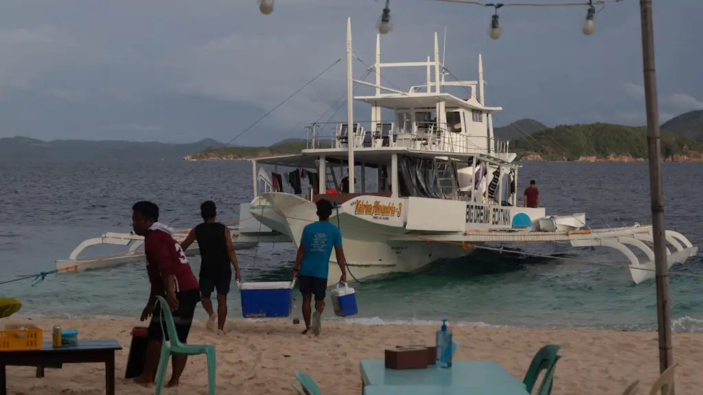 Boat crew carrying cool boxes across a beach towards a white outrigger boat moored in the shallows