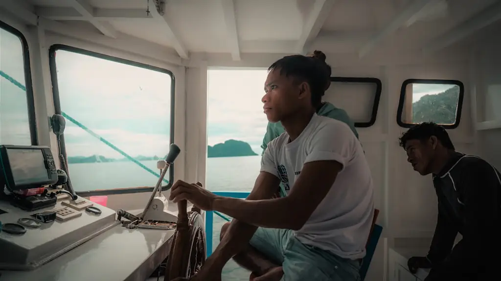 A young crew member steering from a boat wheelhouse with two others beside him and islands visible through the windows