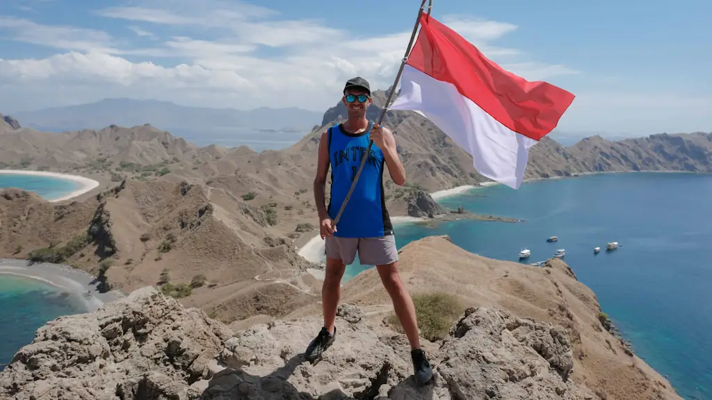 A hiker holding an Indonesian flag on a dry rocky ridge above curved beaches and anchored boats