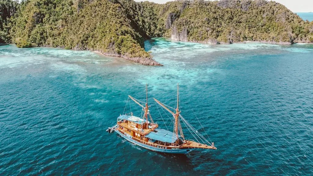 A wooden phinisi at anchor in clear turquoise water beside a forested limestone island